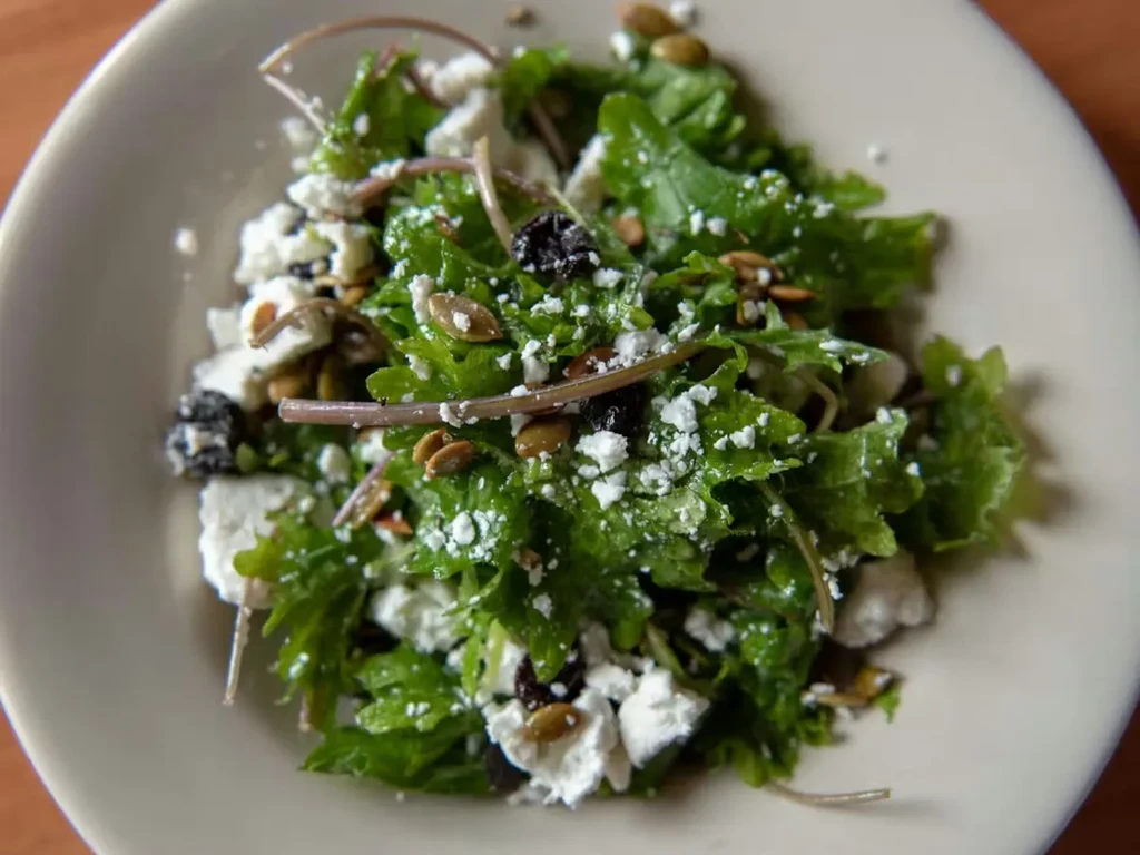 Overhead close-up of green salad, crumbled cheese, dried fruit, pumpkin seeds.