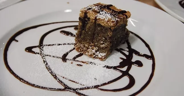 Square dessert with powdered sugar and dark sauce on a white plate.