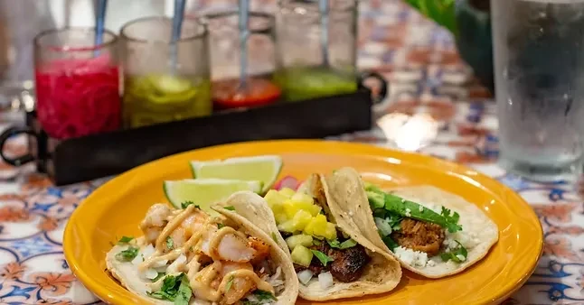 Three distinct tacos on an orange plate with lime and condiments.