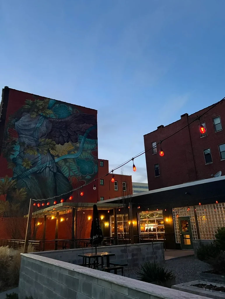 Urban restaurant exterior at twilight with mural, patio, and string lights.