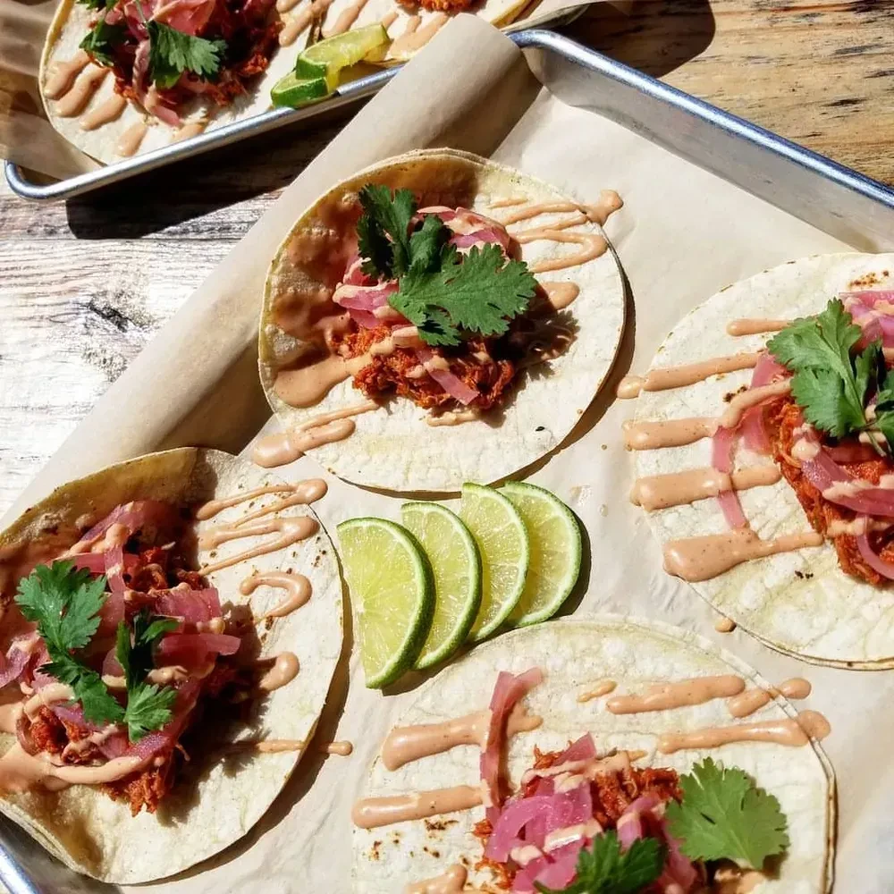 Multiple tacos on baking sheet with meat, pickled onions, cilantro, lime.