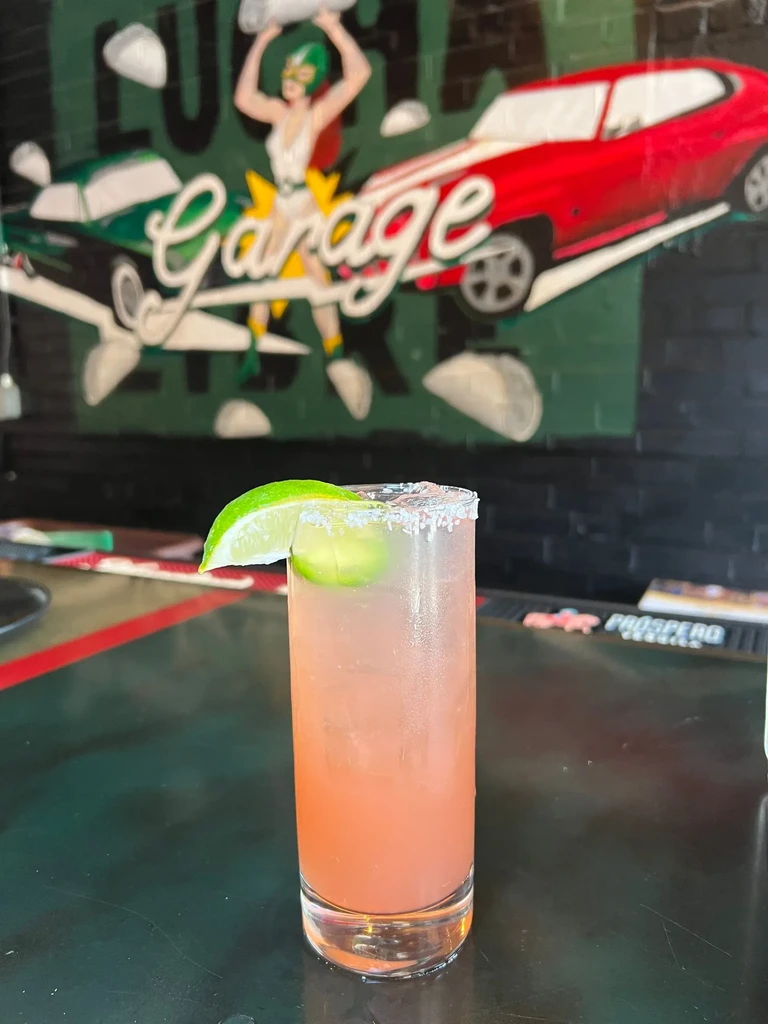 Pinkish-orange cocktail with lime and salt, against a luchador mural.