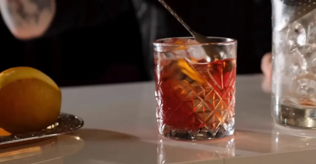 Bartender stirring an amber cocktail in an ornate glass on a bar.