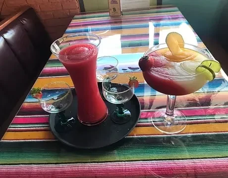 Two vibrant frozen margaritas and small glasses on a striped tablecloth.