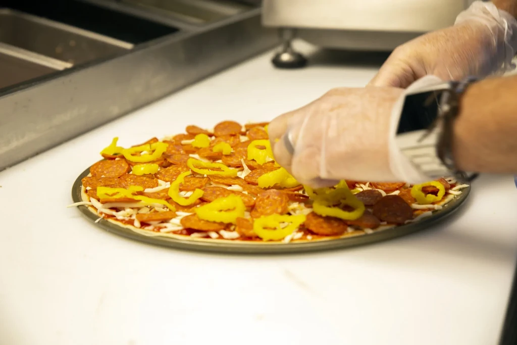 Gloved hands adding yellow peppers to a pepperoni pizza.