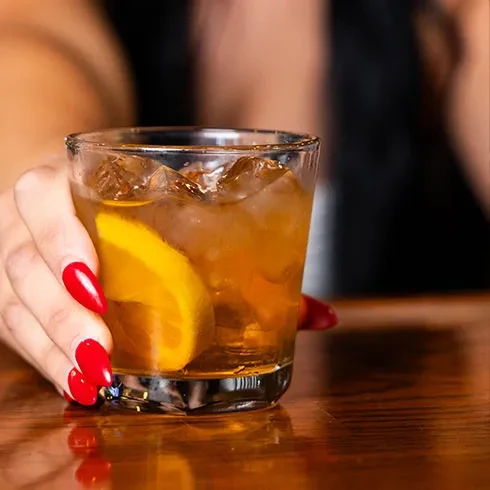 Hand holds amber drink with ice, orange slice, on wood surface.