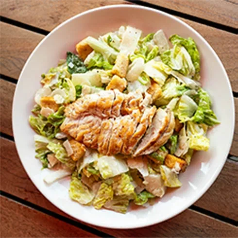 Overhead Caesar salad with sliced grilled chicken in a white bowl.