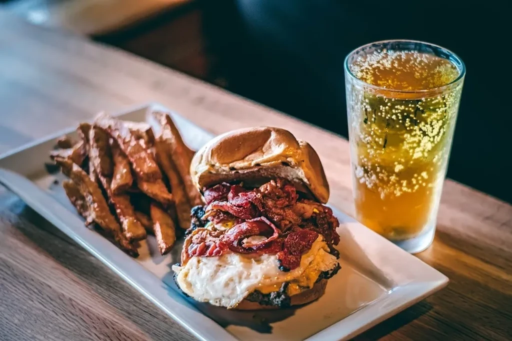 Large burger with egg, bacon, fries, and beer on a wooden table.