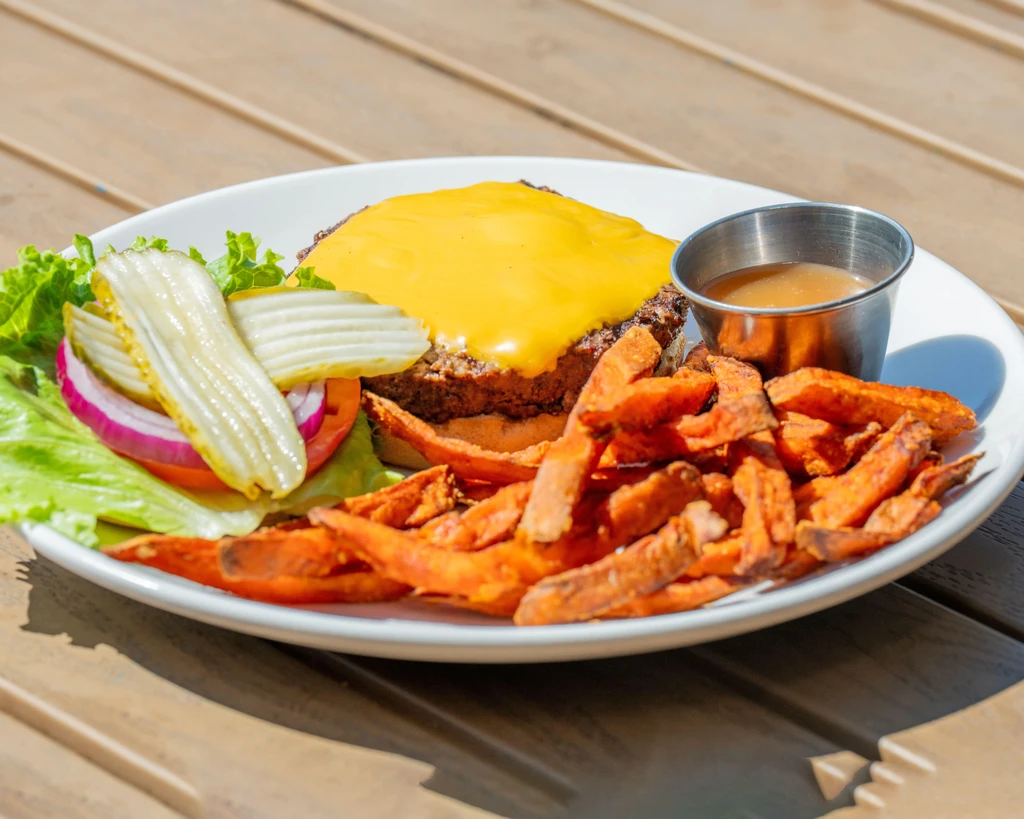 Cheeseburger, sweet potato fries, pickles, and sauce on a white plate outdoors.
