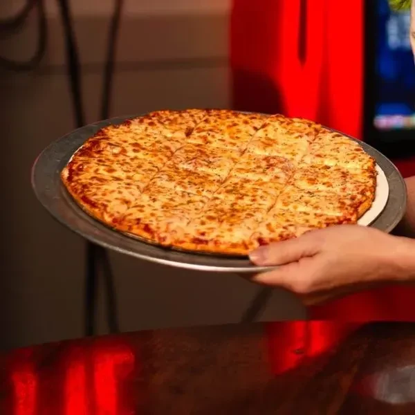 Hands hold round pizza with bubbling cheese, cut into square slices.