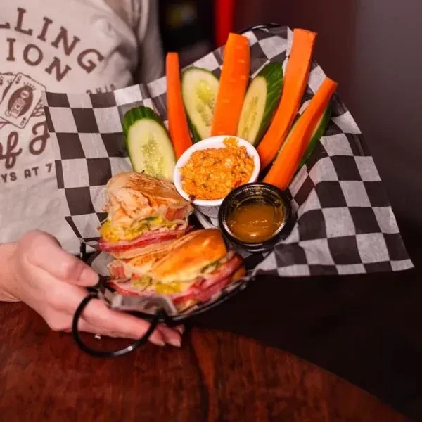 Close-up of held black wire basket with sandwich, vegetables, and dips.