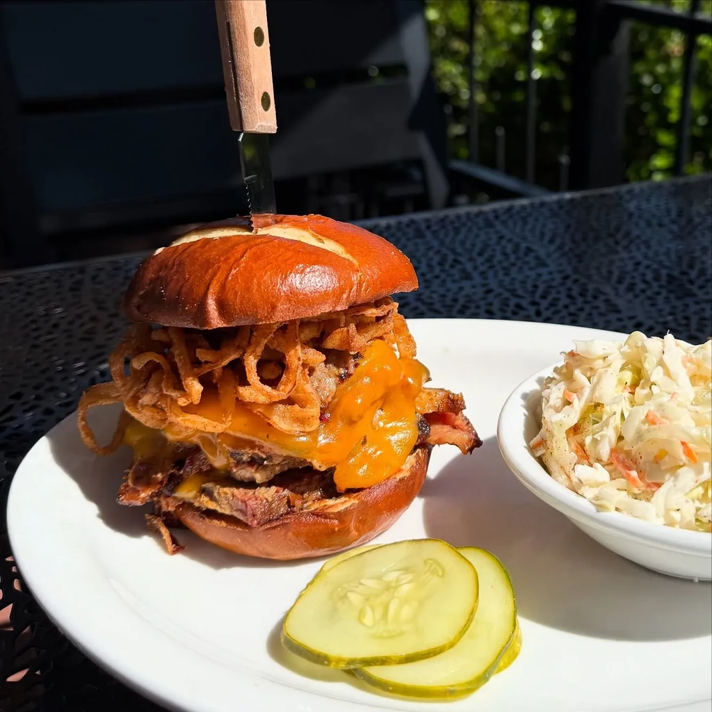 Large pretzel bun sandwich with meat, cheese, fried onions, knife, coleslaw.