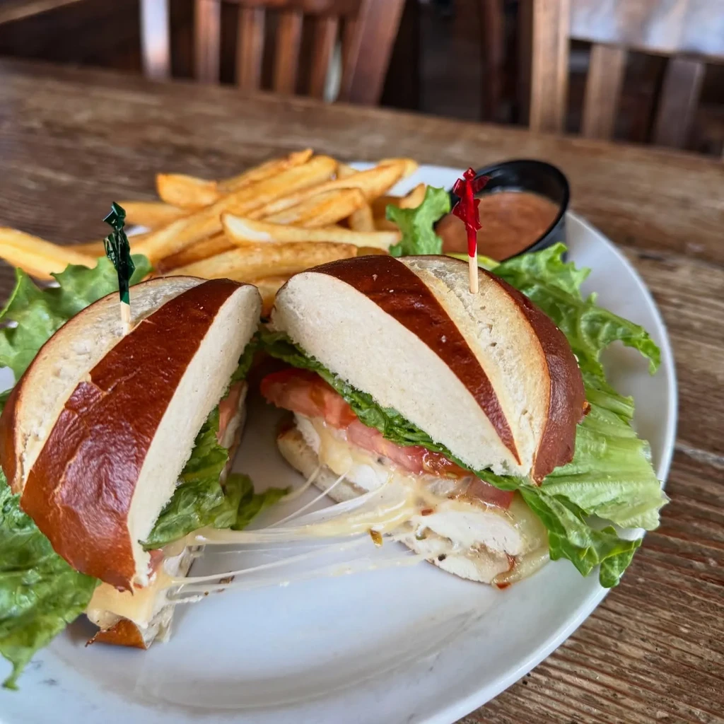 Pretzel bun sandwich with melted cheese, chicken, lettuce, fries, sauce.