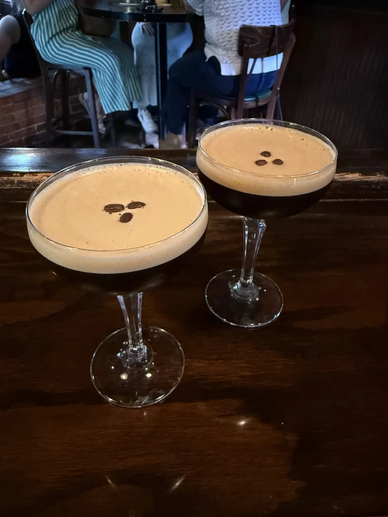 Two espresso martinis with foam and coffee beans on a dark bar.