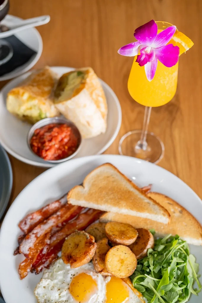 A restaurant table with breakfast plate, burrito, salsa, and a mimosa.