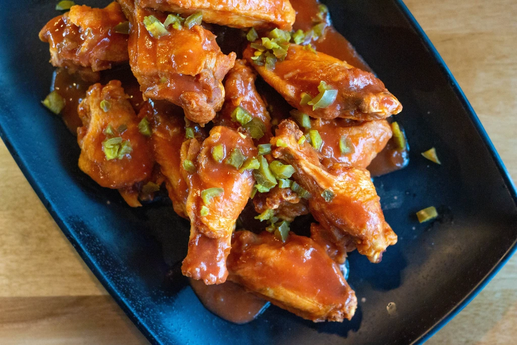 Saucy orange-red chicken wings with green jalapeños on a dark plate.