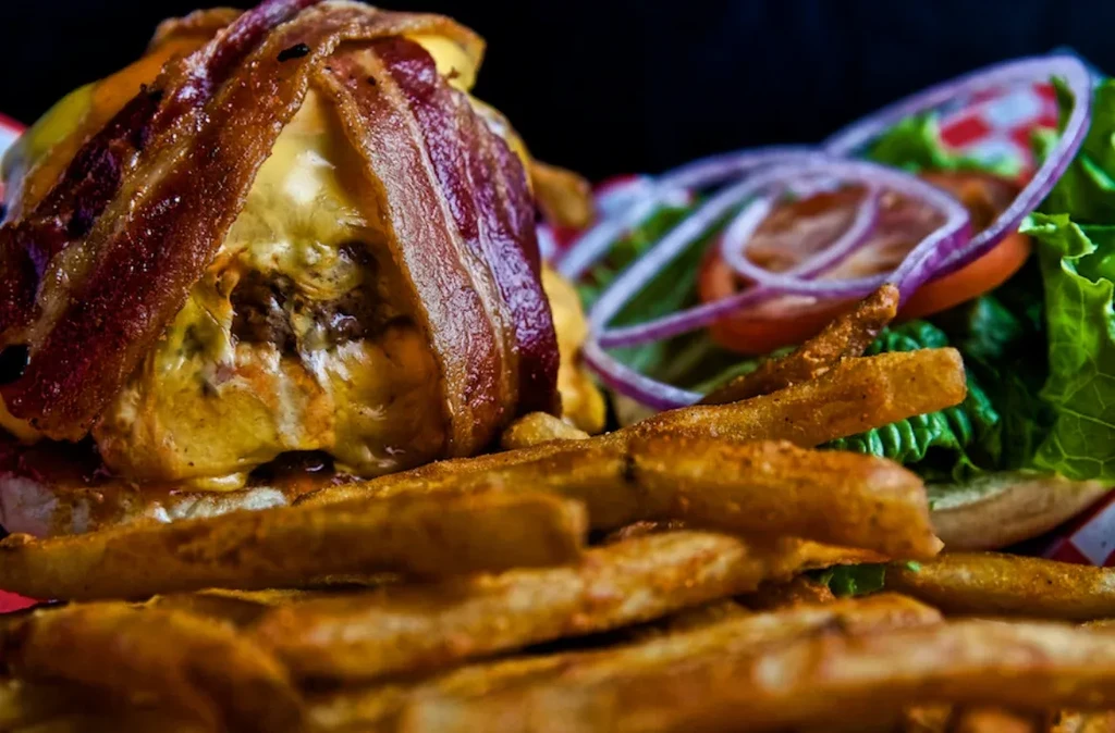Close-up of a bacon-wrapped burger with melted cheese, fries, and salad.