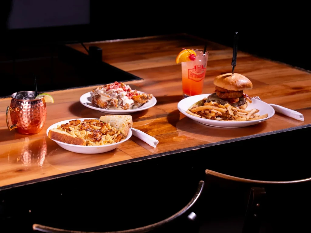 Wooden table with pasta, fried chicken, a burger, and two cocktails.