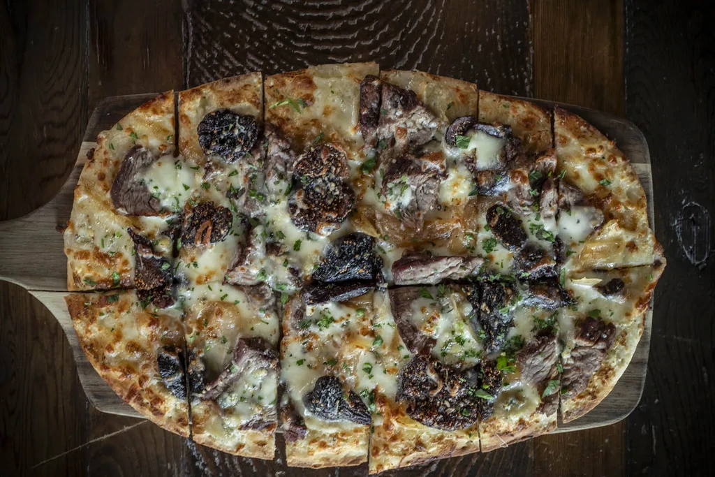 Overhead, oval flatbread with meat, melted cheese, dark fruit, and herbs.