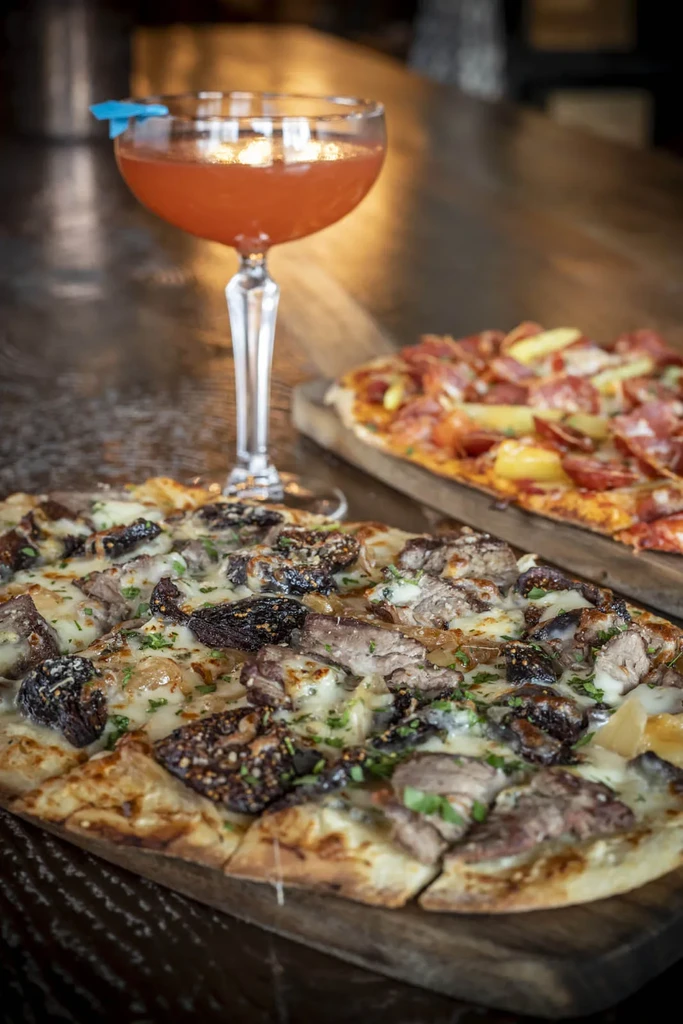 Fig and meat pizza, pepperoni pizza, and airplane cocktail on dark table.