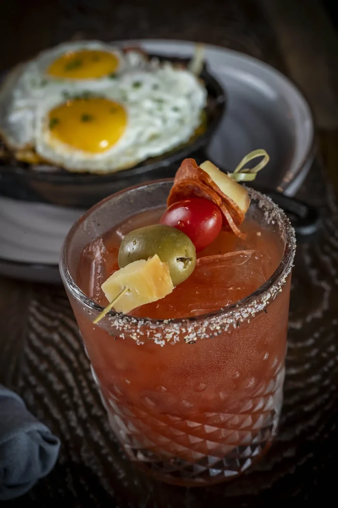 Garnished Bloody Mary-style cocktail, with blurred sunny-side-up eggs.