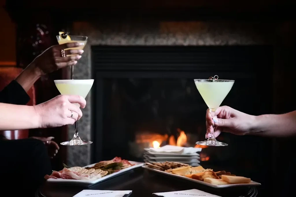 Hands holding fruit-garnished cocktails and appetizers by a warm fireplace.