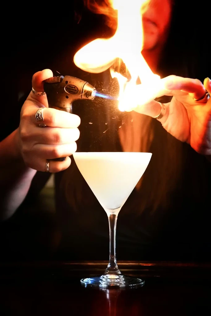 Hands torching a cocktail, producing large orange and yellow flames.