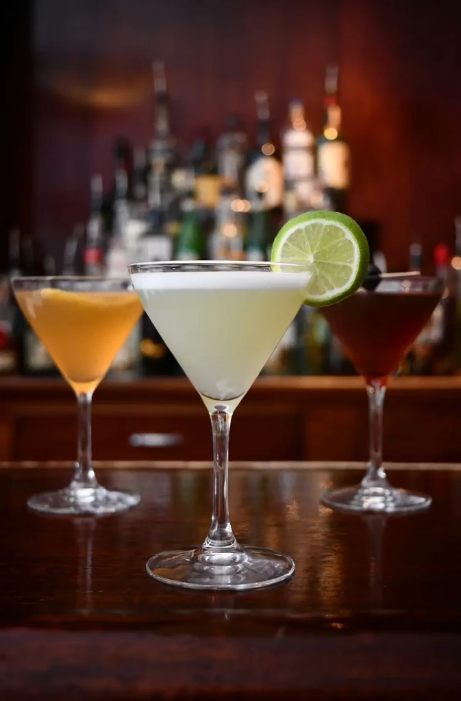 Three cocktails in martini glasses, one with lime, on a wooden bar.