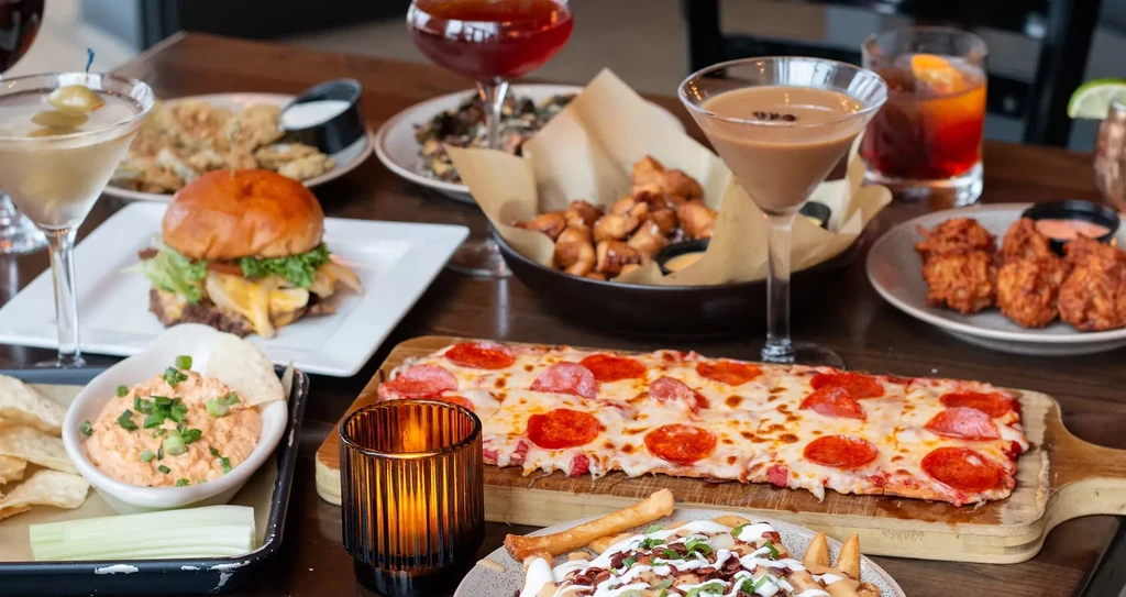 Flat-lay of assorted cocktails, appetizers, hamburger, pizza, and loaded fries.