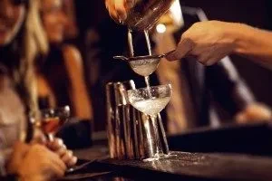 Bartender pouring frothing white cocktail from shaker into coupe glass.