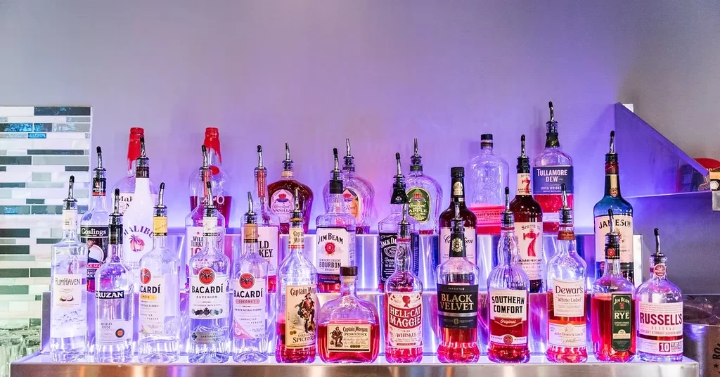 Well-stocked bar shelf with diverse liquor bottles and colorful background lighting.