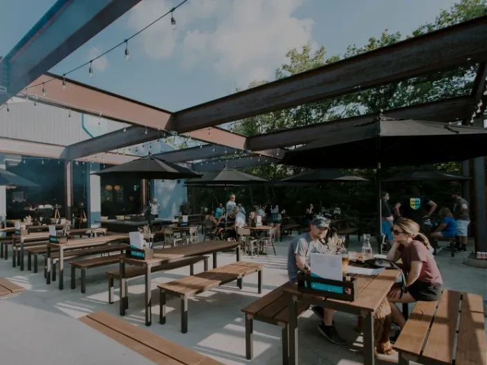 Rustic outdoor patio with picnic tables, umbrellas, string lights, and guests.