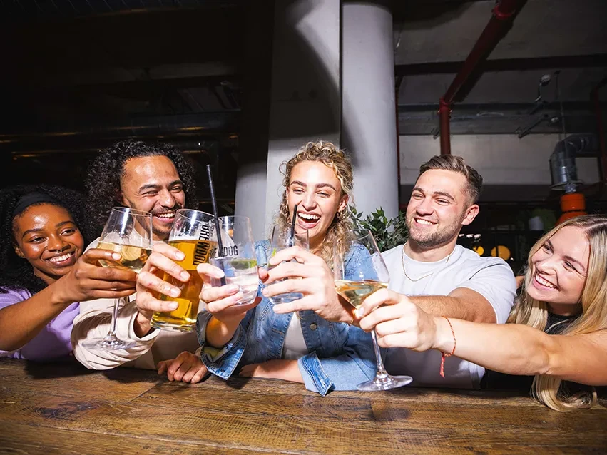 Five diverse young adults smiling, toasting drinks in a bar.