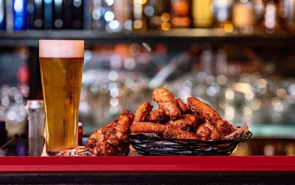Beer glass, basket of golden-brown chicken wings on a bar.