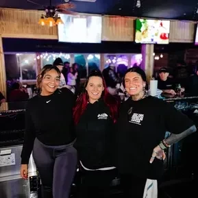 Three smiling bar staff women stand in front of a counter.