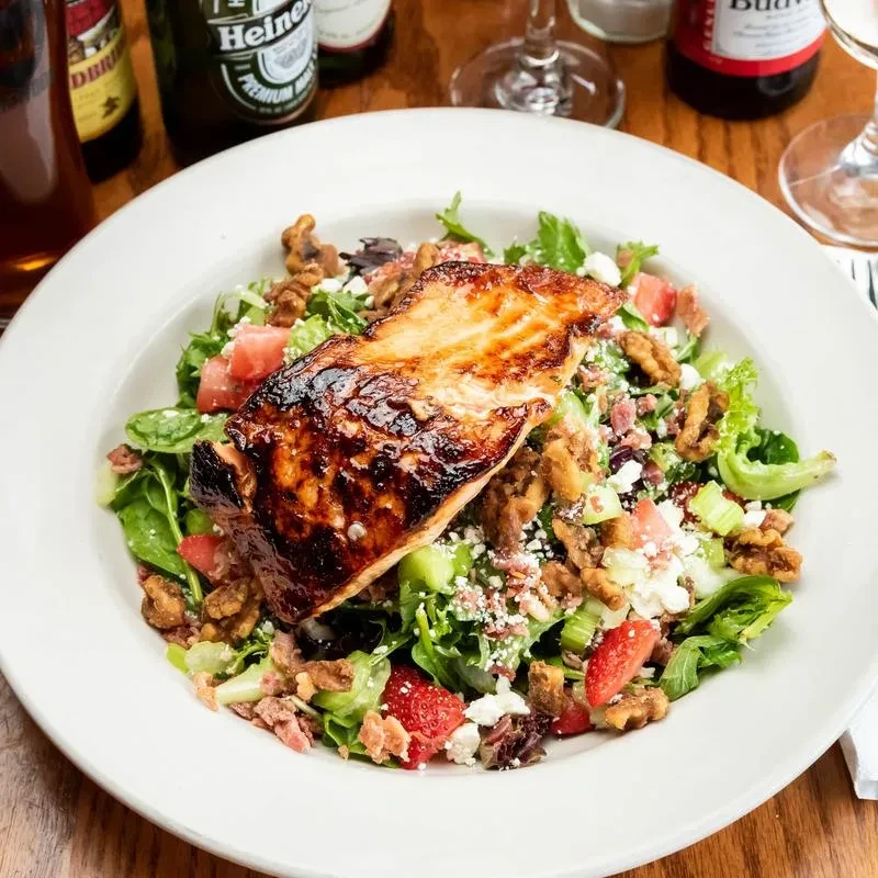 Grilled salmon salad in a white bowl with greens, strawberries, and walnuts.