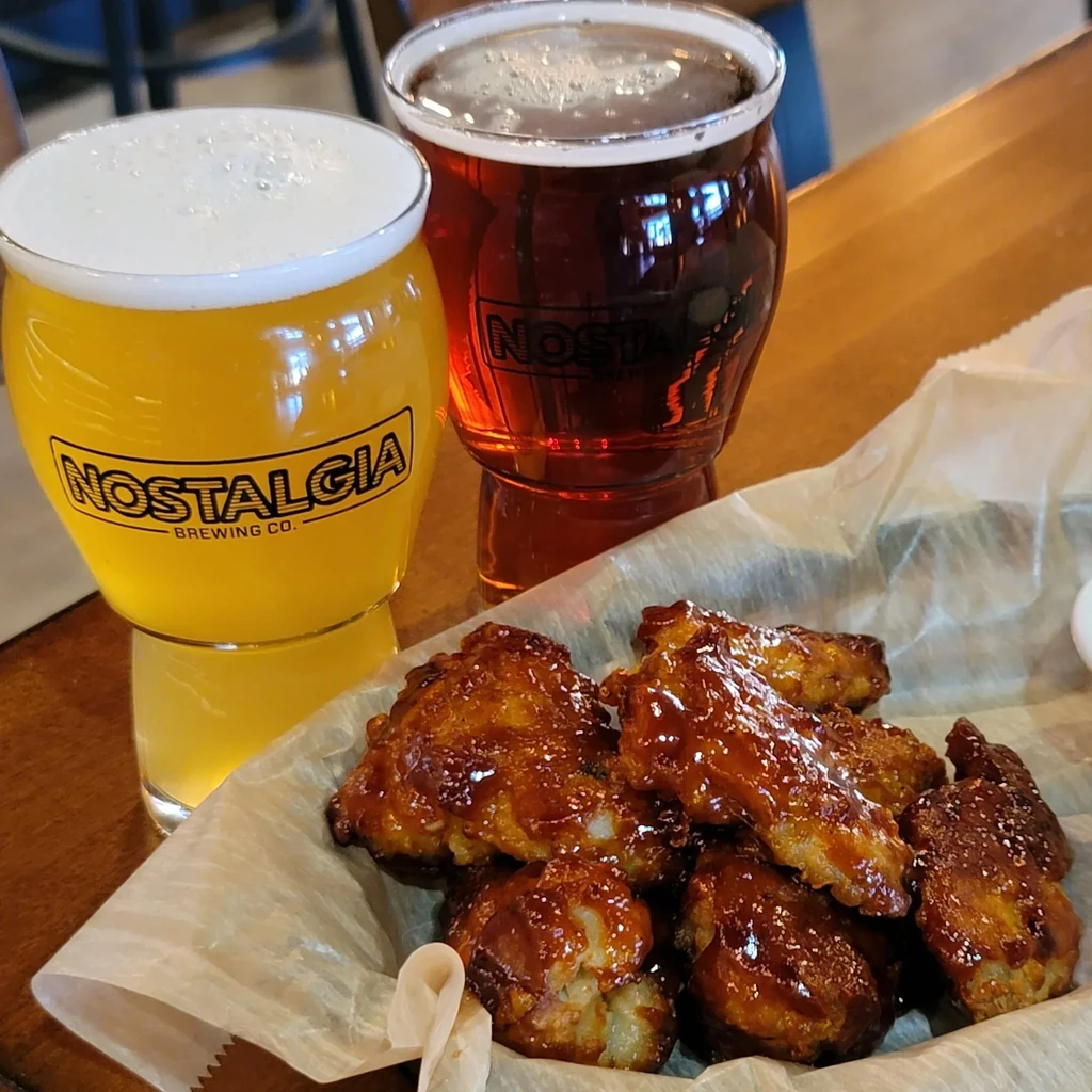 Two Nostalgia Brewing beers and chicken wings on a wooden table.