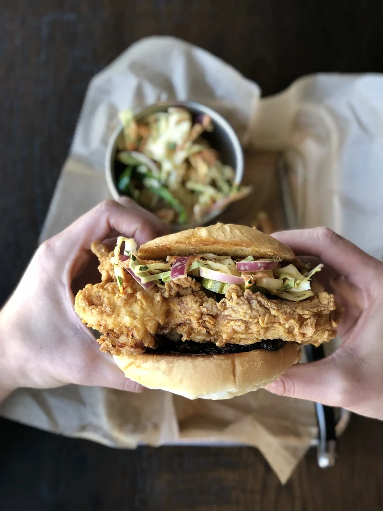 Large fried chicken sandwich with coleslaw and pickles, held by hands.