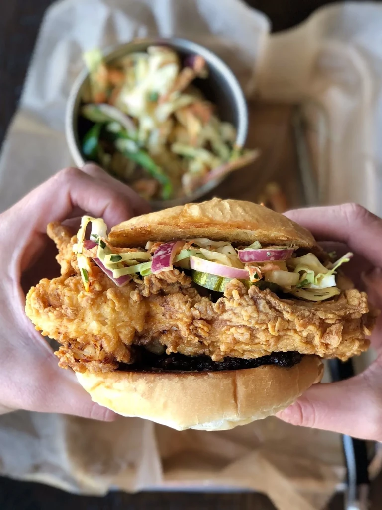 Large fried chicken sandwich topped with colorful coleslaw, held by hands.