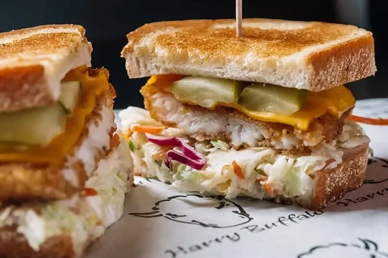Halved fried fish sandwich on toasted bread with cheese, pickles, coleslaw.