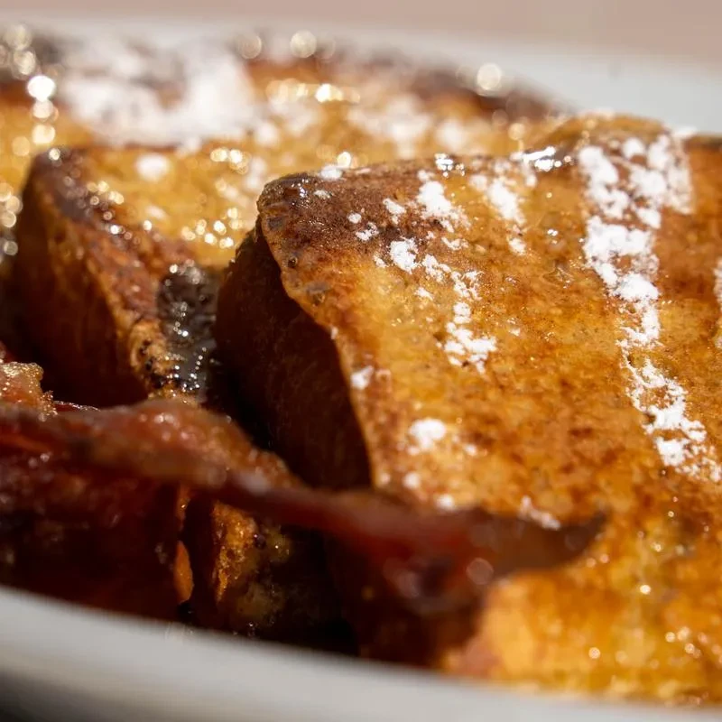 Plate of golden French toast with powdered sugar, maple syrup, and bacon.