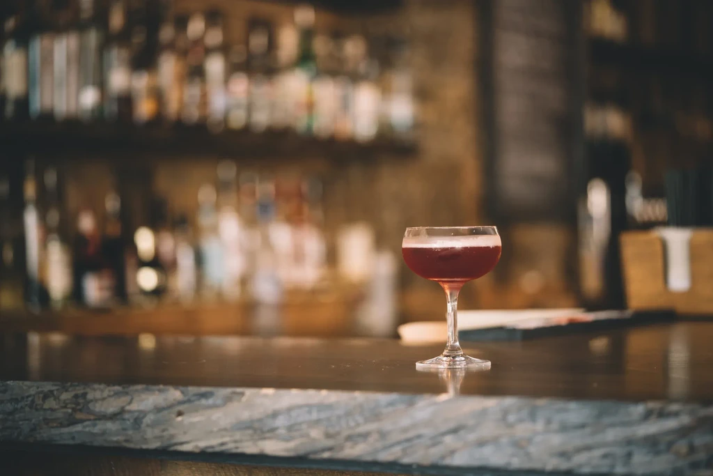 Dark red cocktail in a coupe glass on a dimly lit bar.