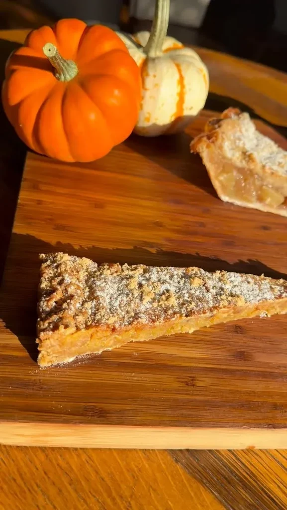 Two slices of fruit crumble pie with powdered sugar on wood with pumpkins.