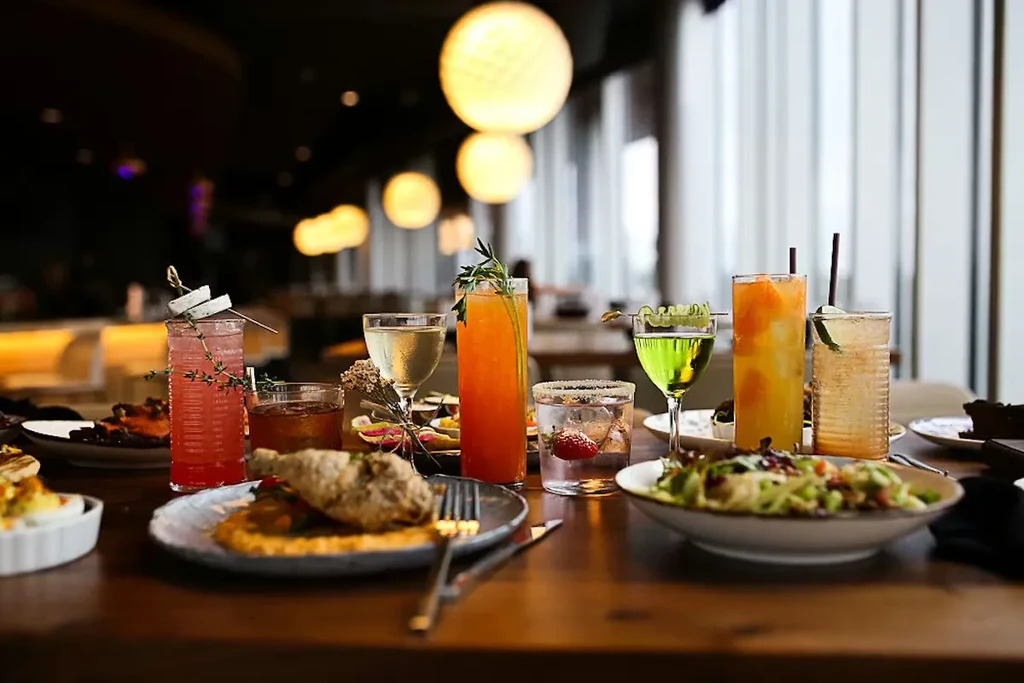 Seven colorful cocktails, fried chicken, and salad on a restaurant table.