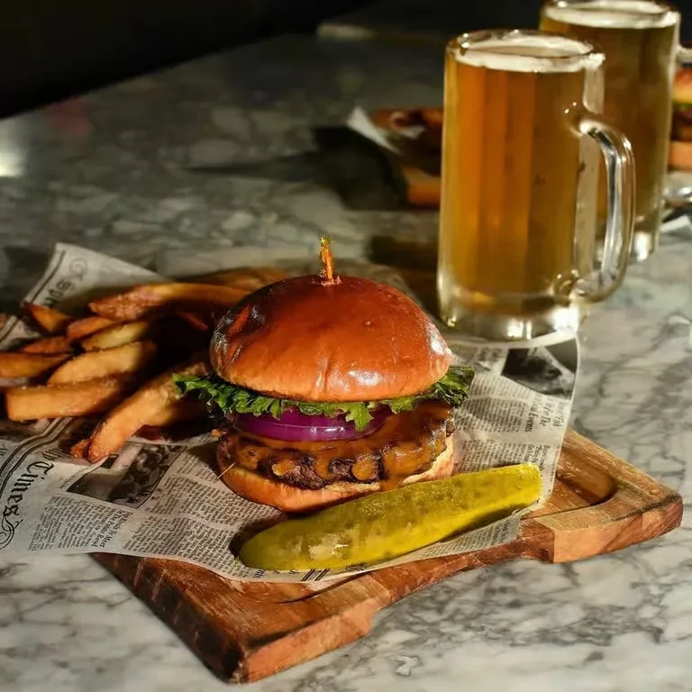 Gourmet burger, fries, pickle, and two beers on a wooden board.