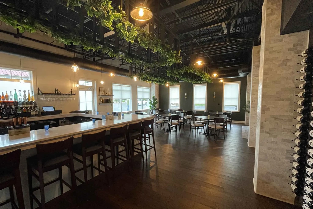 Modern restaurant interior with long bar, dining, exposed beams, wine rack.