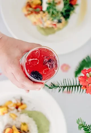 Overhead: Hand holding red berry drink with blackberries, ice, and food.