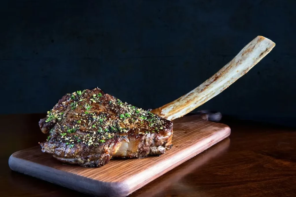 Seared tomahawk steak with bone, garnished with herbs on a wood board.