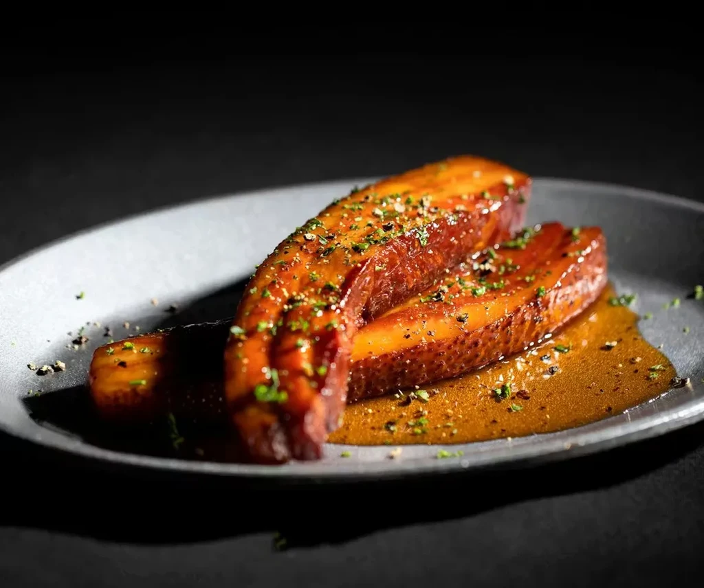 Two glazed pork belly pieces with herbs and pepper on a grey plate.