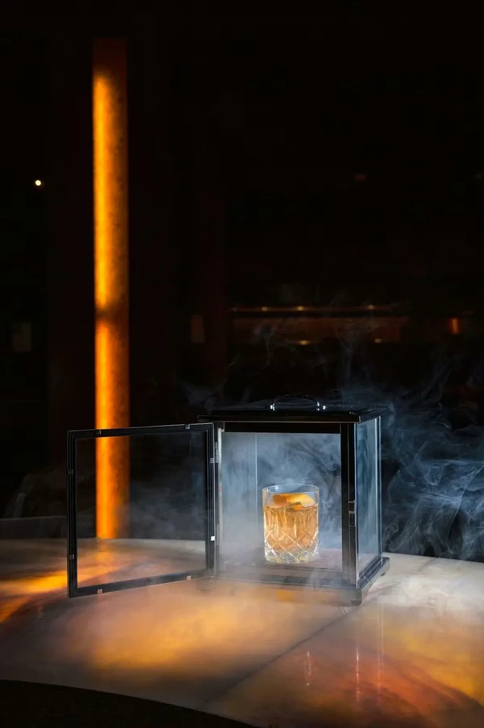 Cocktail with orange peel in cut-glass cloche, smoke billowing out.
